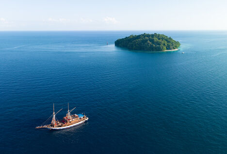 Aerial view of a tropical island, surrounded by coral reef with a liveaboard sailing boat