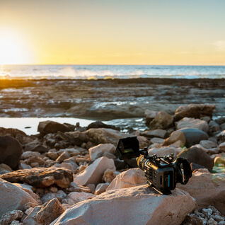 A large camera sits on a rock on the shoreline of an ocean during sunset.