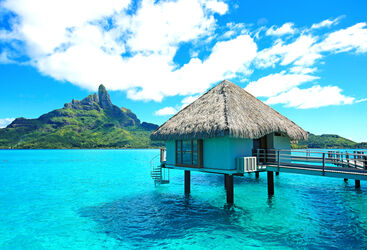 a thathed roof bungalow on silts over clear blue waters with a mountain in the background in Bora Bora