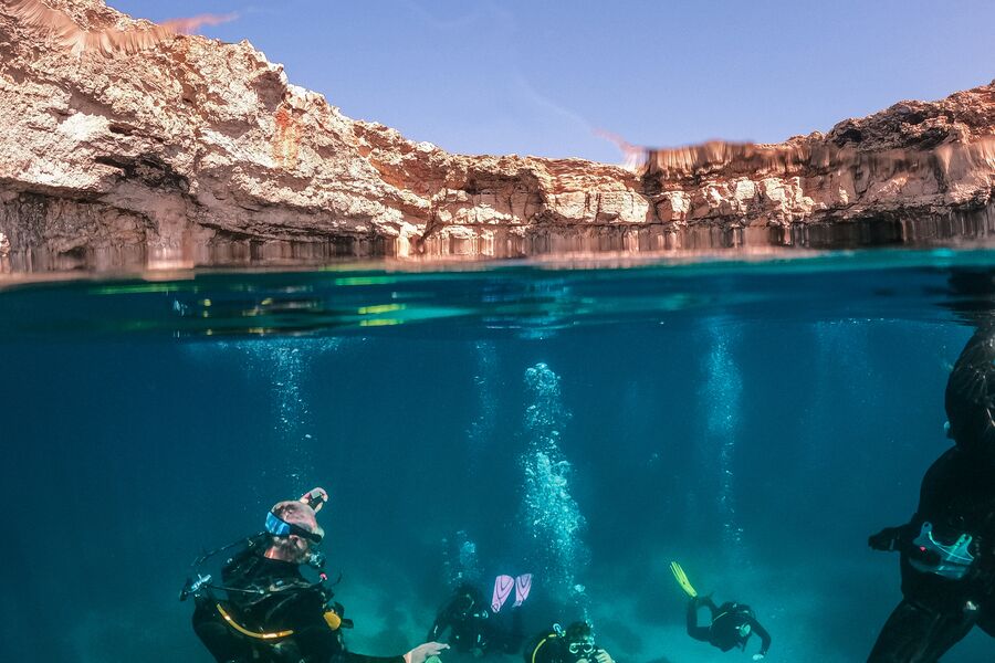 Split view water level photo of divers descending into clear blue waters with sand stone aurrounding them above the surface.