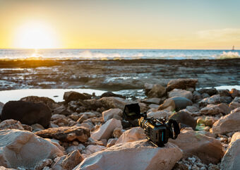 A large camera sits on a rock on the shoreline of an ocean during sunset.